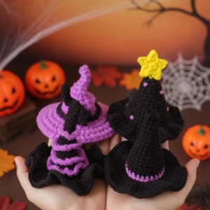 Può includere: Due cappelli da strega all'uncinetto fatti a mano. Uno è viola e nero, l'altro è nero con una stella gialla. I cappelli sono esposti su uno sfondo a tema Halloween con zucche e una ragnatela.