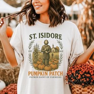 May include: Cream-colored t-shirt with a graphic that reads "St. Isidore Pumpkin Patch Patron Saint of Farmers." The shirt is paired with a pumpkin spice baseball cap. Pumpkins and hay bales are in the background.