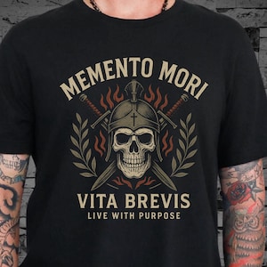 Puede incluir: Camiseta negra con un dise&ntilde;o de calavera con casco y espadas cruzadas. El texto "Memento Mori" est&aacute; sobre la calavera y "Vita Brevis Live With Purpose" debajo. El dise&ntilde;o incluye ramas de olivo y llamas.