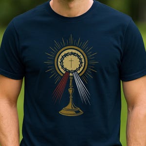 May include: Navy blue t-shirt featuring a gold-colored religious symbol with a cross, crown of thorns, and rays of light. The design includes red and white vertical lines. The shirt is made of a soft material.
