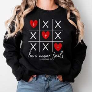 Könnte beinhalten: Schwarzes Sweatshirt mit einem Tic-Tac-Toe-Design mit roten Herzen und Kreuzen. Der Schriftzug "love never fails" steht in Kursivschrift unter dem Spielbrett, darunter der Bibelvers 1. Korinther 13,8.