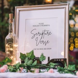 May include: A white sign with a black frame, sitting on a table with greenery. The sign says "Please highlight a meaningful Scripture Verse for the bride and groom".