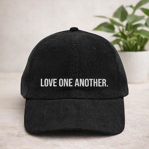 Op de afbeelding: Zwarte baseballcap met de witte tekst "LOVE ONE ANOTHER." De cap is gemaakt van een textuurstof en heeft een gebogen klep. Een klassieke stijl cap, geschikt voor casual wear.