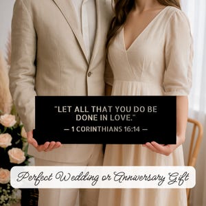 Op de afbeelding: Een zwart rechthoekig bord met de tekst "LET ALL THAT YOU DO BE DONE IN LOVE." - 1 CORINTHIANS 16:14 -" vastgehouden door een stel. Het bord bevat ook de tekst "Perfect Wedding or Anniversary Gift". Het paar draagt formele kleding.