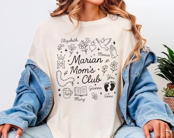 May include: Cream-colored t-shirt with the text "Marian Mom's Club" in black, surrounded by religious symbols and names. The design includes a crown, flowers, a dove, and a rosary. The shirt is worn with a denim jacket.