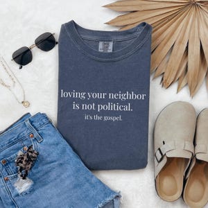 May include: A navy blue sweatshirt with the text "loving your neighbor is not political. it's the gospel." is displayed. The sweatshirt is folded next to denim shorts, sunglasses, a necklace, and sandals.