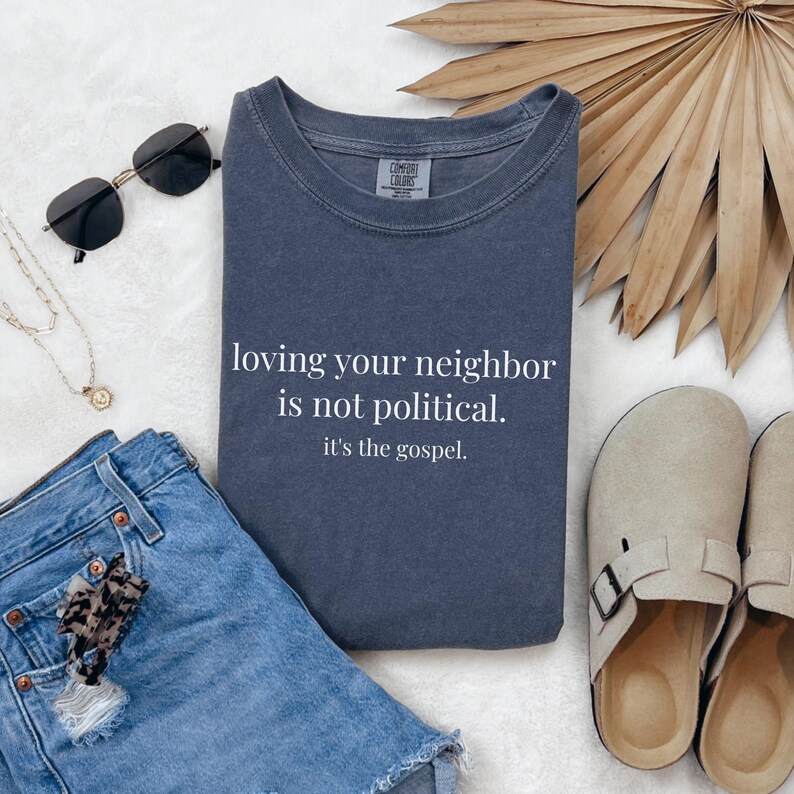 May include: A navy blue sweatshirt with the text "loving your neighbor is not political. it's the gospel." is displayed. The sweatshirt is folded next to denim shorts, sunglasses, a necklace, and sandals.