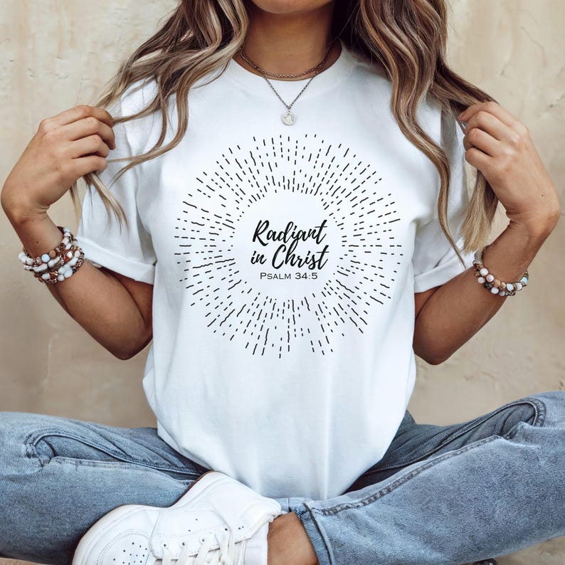 May include: White t-shirt with a black sunburst design and the text "Radiant in Christ" with the scripture reference "Psalm 34:5".