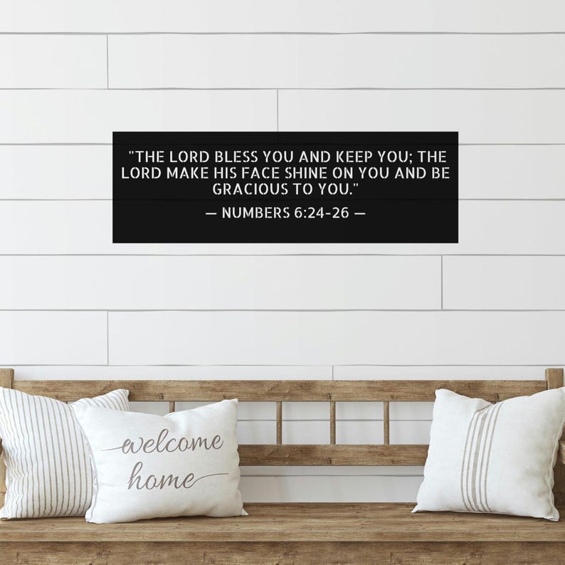 Op de afbeelding: Een zwart houten bord met de tekst "THE LORD BLESS YOU AND KEEP YOU; THE LORD MAKE HIS FACE SHINE ON YOU AND BE GRACIOUS TO YOU." uit Numeri 6:24-26. Het bord hangt boven een houten bank met kussens.