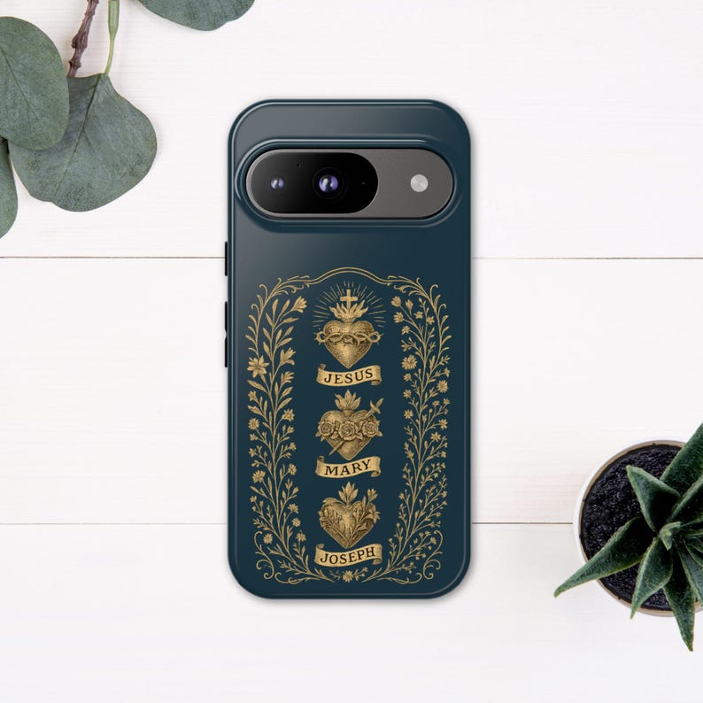 May include: A dark teal phone case featuring a gold illustration of three hearts labeled "Jesus," "Mary," and "Joseph." The design is framed by a gold floral border. The phone case is on a white wooden surface with a small potted plant.