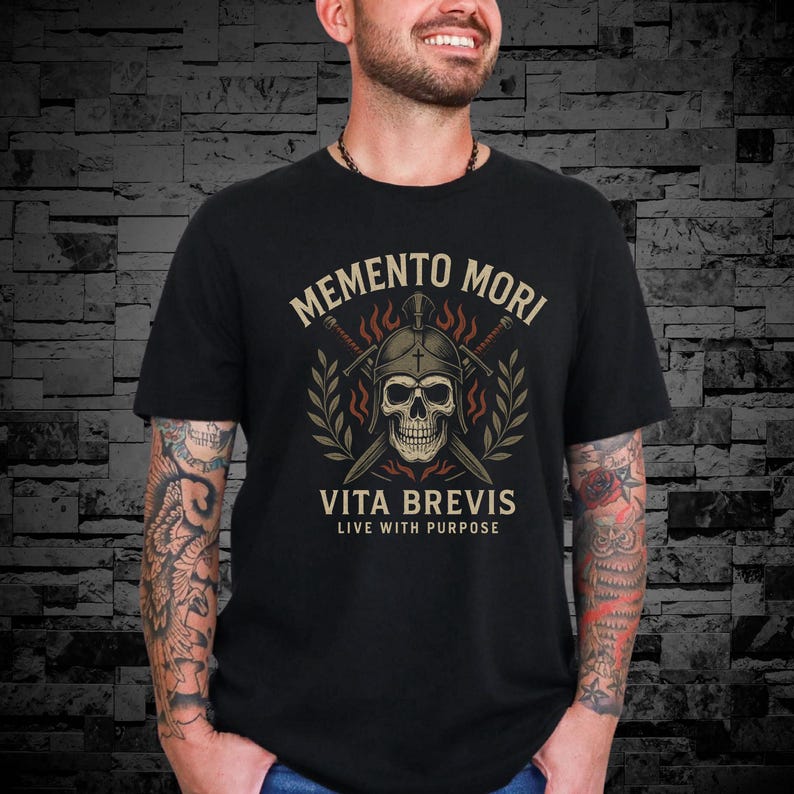 Puede incluir: Camiseta negra con un dise&ntilde;o de calavera con casco y espadas cruzadas. El texto "Memento Mori" est&aacute; sobre la calavera y "Vita Brevis Live With Purpose" debajo. El dise&ntilde;o incluye ramas de olivo y llamas.