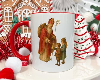 May include: White ceramic mug with a red handle and rim. The mug features a vintage illustration of Saint Nicholas giving a doll to two children. The saint is dressed in a red robe and carries a staff and a sack. Festive holiday decor surrounds the mug.