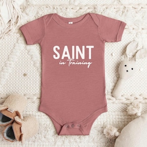 May include: A dusty rose baby onesie with short sleeves and white text that reads "SAINT in TRAINING".