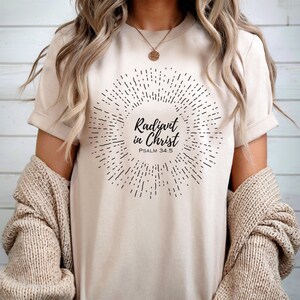 May include: A beige t-shirt with a black graphic design of a sunburst with the text "Radiant in Christ" and "Psalm 34:5".