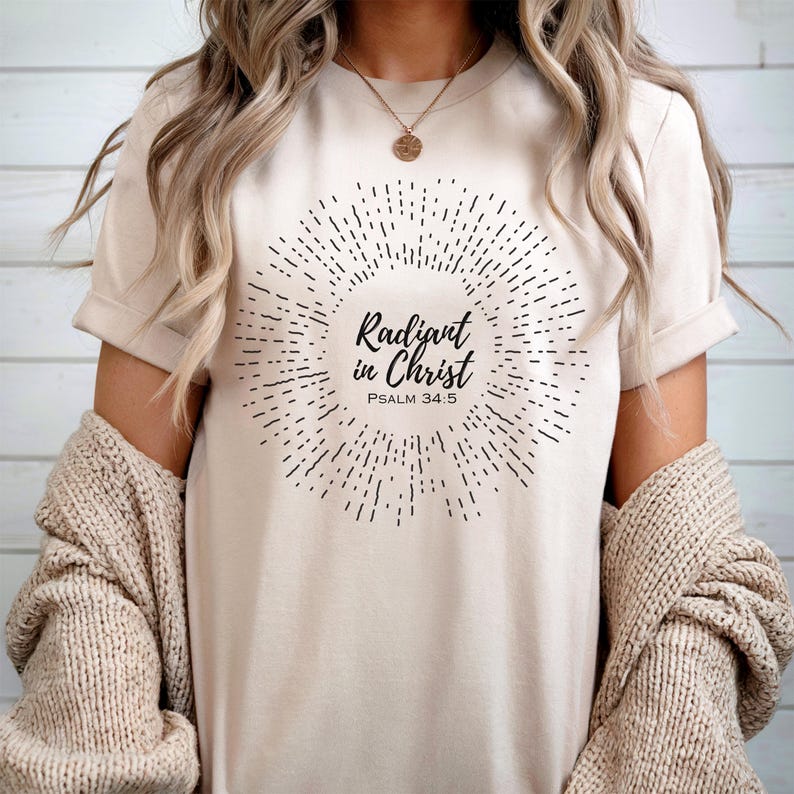 May include: A beige t-shirt with a black graphic design of a sunburst with the text "Radiant in Christ" and "Psalm 34:5".