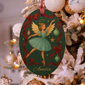 May include: Oval Christmas ornament featuring a ballerina angel in a teal tutu and gold wings, with the name "Amelia" at the bottom. The dark green background is decorated with red ribbons, bows, and ornaments. A red ribbon is attached for hanging.