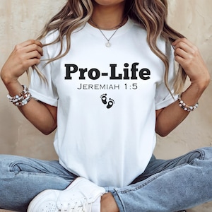 Puede incluir: Camiseta blanca con texto negro que dice "Pro-Life" y "Jeremías 1:5". La camiseta también presenta un par de pequeñas huellas negras.