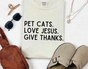 May include: Cream-colored t-shirt with the text "PET CATS. LOVE JESUS. GIVE THANKS." Black sunglasses, a gold hair clip, a brown crossbody bag, a gold necklace, and beige slip-on shoes are arranged around the shirt on a white textured surface.