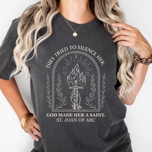 May include: Dark grey t-shirt with the text "THEY TRIED TO SILENCE HER" and an illustration of a sword and flame. The text "GOD MADE HER A SAINT. ST. JOAN OF ARC" is below the illustration.