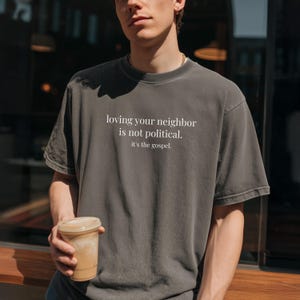 May include: Dark gray t-shirt with the text "loving your neighbor is not political. it's the gospel." in white. The shirt has a vintage, slightly faded look. The shirt is a crew neck style.