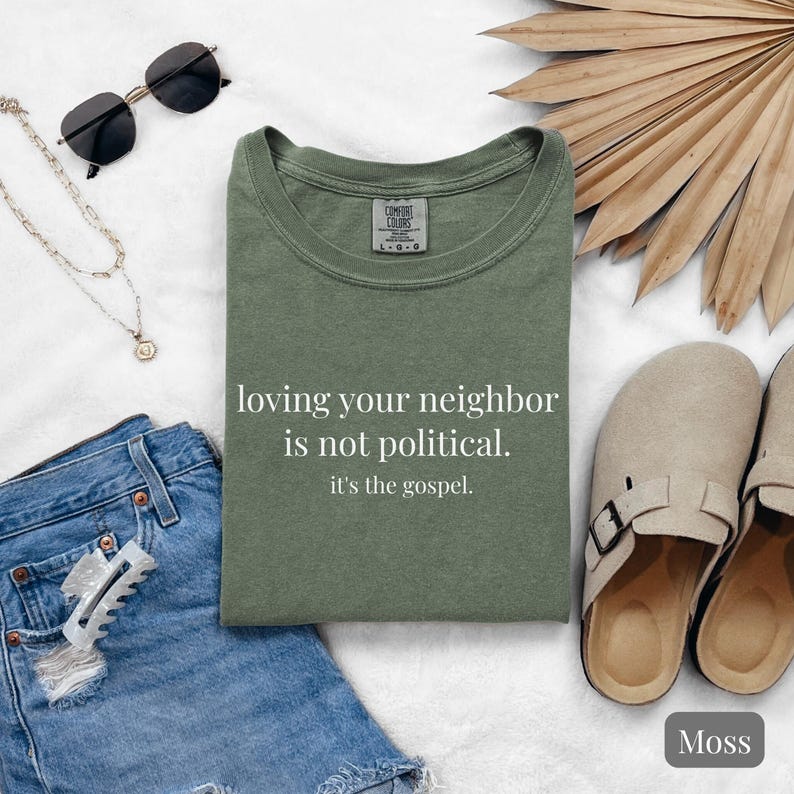 May include: An olive green t-shirt with the text "loving your neighbor is not political. it's the gospel." is displayed. The shirt is folded and surrounded by accessories, including sunglasses, a necklace, denim shorts, a hair clip, and sandals. The shirt is labeled "Moss".