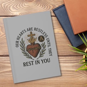May include: A grey journal featuring the text "OUR HEARTS ARE RESTLESS UNTIL THEY REST IN YOU" and a heart, cross, and flame graphic. The journal is on a wooden surface with other books and a plant.
