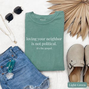 May include: Light green t-shirt with the text "loving your neighbor is not political. it's the gospel." displayed on a white background. The shirt is paired with denim shorts, sunglasses, a necklace, and sandals.