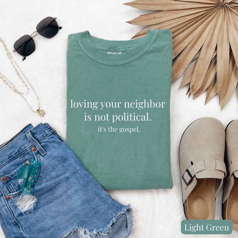 May include: Light green t-shirt with the text "loving your neighbor is not political. it's the gospel." displayed on a white background. The shirt is paired with denim shorts, sunglasses, a necklace, and sandals.