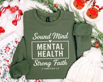 May include: An olive green sweatshirt with the words "Sound Mind, Mental Health, Strong Faith" in white. The sweatshirt is surrounded by holiday decorations, including a pair of reindeer slippers, a white knit hat, and festive greenery.