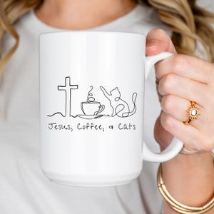 May include: White ceramic mug with a black line drawing of a cross, coffee cup, and cat. The text "Jesus, Coffee, & Cats" is below the drawing. The mug is held by a person.
