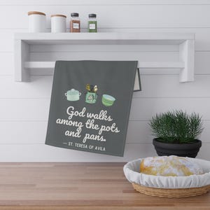 Könnte beinhalten: Ein dunkelgraues Küchentuch mit dem Zitat „Gott wandelt unter Töpfen und Pfannen“ und Illustrationen von Küchenutensilien. Das Tuch hängt an einem weißen Regal über einer Holzoberfläche.