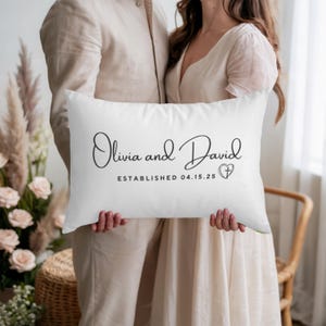 Puede incluir: Un cojín rectangular blanco con los nombres "Olivia and David" en escritura cursiva, la fecha "04.15.25" y un símbolo de corazón. El cojín es sostenido por dos personas.