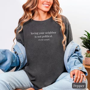 May include: A dark gray sweatshirt with the text "loving your neighbor is not political. it's the gospel." in white. The sweatshirt is part of a casual outfit, paired with a denim jacket and jeans.
