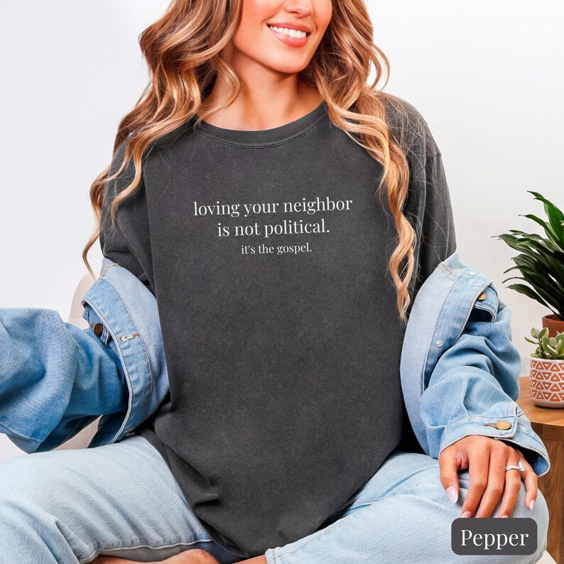 May include: A dark gray sweatshirt with the text "loving your neighbor is not political. it's the gospel." in white. The sweatshirt is part of a casual outfit, paired with a denim jacket and jeans.