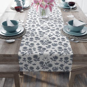 May include: A white table runner featuring a repeating pattern of dark blue roses, crowns, and other decorative motifs. The runner is laid on a wooden table set for a meal, with plates, bowls, and glasses.