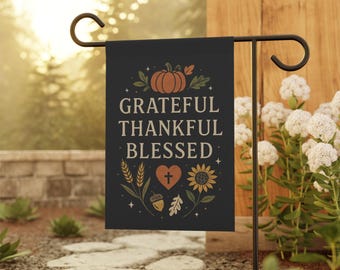 May include: A black garden flag with the words "Grateful, Thankful, Blessed" in white. The flag features a pumpkin, wheat, a heart with a cross, a sunflower, an acorn, and leaves in shades of orange and yellow.