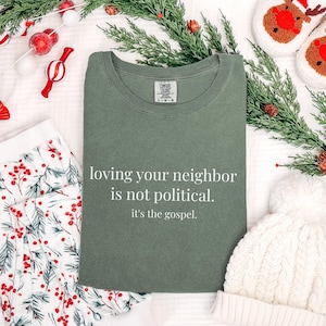 May include: An olive green t-shirt with the text "loving your neighbor is not political. it's the gospel." is displayed. The shirt is surrounded by holiday decorations, including greenery, a white beanie, and festive pajamas.