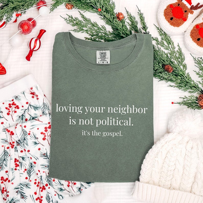 May include: An olive green t-shirt with the text "loving your neighbor is not political. it's the gospel." is displayed. The shirt is surrounded by holiday decorations, including greenery, a white beanie, and festive pajamas.