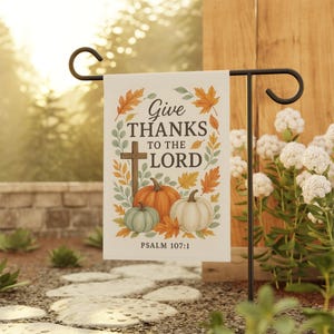 Könnte beinhalten: Eine Gartenflagge mit dem Text "Give Thanks to the Lord" und Psalm 107:1. Die Flagge zeigt ein Kreuz, Kürbisse und Herbstblätter in Orange, Grün und Weiß. Sie wird auf einem schwarzen Metallständer in einem Garten gezeigt.