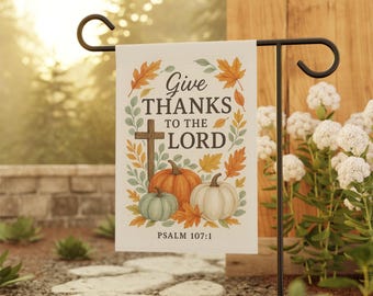 May include: A garden flag with the text "Give Thanks to the Lord" and Psalm 107:1. The flag features a cross, pumpkins, and autumn leaves in orange, green, and white. It is displayed on a black metal stand in a garden setting.
