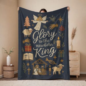 May include: A navy blue blanket with a Christmas theme. The blanket features illustrations of an angel, a sheep, a Christmas tree, and the text "Glory to the newborn King". Other illustrations include a candle, a star, and a gift.