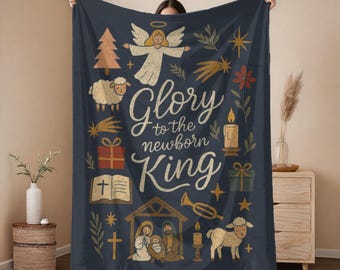 Puede incluir: Una manta azul marino con temática navideña. La manta presenta ilustraciones de un ángel, una oveja, un árbol de Navidad y el texto "Gloria al Rey recién nacido". Otras ilustraciones incluyen una vela, una estrella y un regalo.