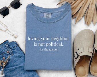 May include: A blue jean colored t-shirt with the text "loving your neighbor is not political. it's the gospel." The shirt is folded and surrounded by accessories including sunglasses, jewelry, and denim shorts.