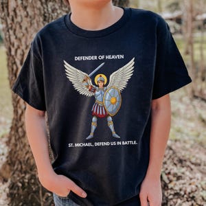 May include: Black t-shirt featuring a graphic of St. Michael, a winged figure in armor holding a sword and shield. The text "DEFENDER OF HEAVEN" and "ST. MICHAEL, DEFEND US IN BATTLE" are printed above and below the illustration.