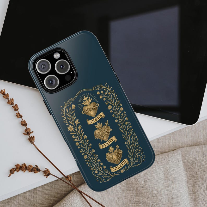 May include: A teal phone case featuring a gold design with three sacred hearts, each labeled with a name: Jesus, Mary, and Joseph. The design is framed by a gold floral border. The phone case is resting on a light-colored surface.