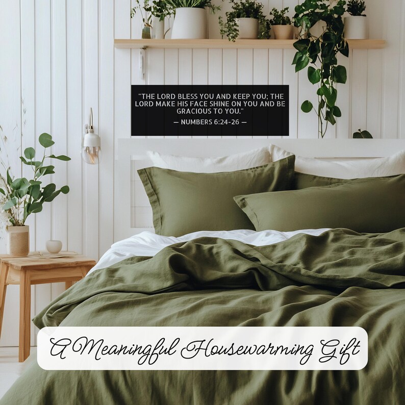 Op de afbeelding: Een slaapkamer met een bed bedekt met olijfgroen beddengoed en kussens. Een zwart bord boven het bed toont een bijbelvers. De tekst "A Meaningful Housewarming Gift" staat onderaan de afbeelding.