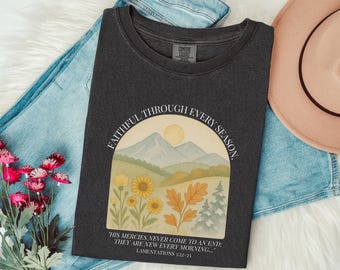 May include: Dark gray t-shirt with a watercolor design featuring a mountain landscape, sunflowers, and the text "FAITHFUL THROUGH EVERY SEASON." The quote "HIS MERCIES NEVER COME TO AN END: THEY ARE NEW EVERY MORNING..." is also included. The shirt is displayed with jeans and a hat.