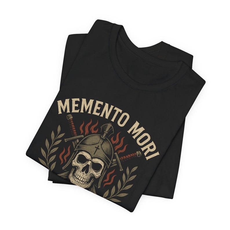 Puede incluir: Camiseta negra con una calavera con casco, espadas cruzadas y las palabras "Memento Mori" en un estilo vintage. El dise&ntilde;o incluye llamas y ramas de laurel. La camiseta est&aacute; doblada.