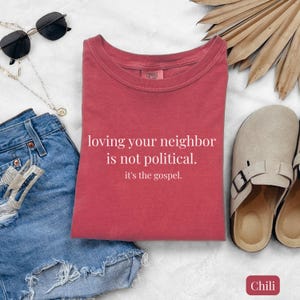 May include: A red t-shirt with the text "loving your neighbor is not political. it's the gospel." is displayed on a white surface. The shirt is paired with denim shorts, sunglasses, a necklace, and beige sandals.