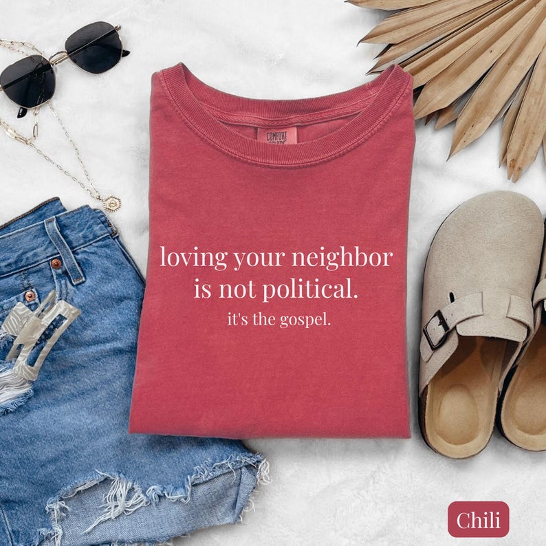 May include: A red t-shirt with the text "loving your neighbor is not political. it's the gospel." is displayed on a white surface. The shirt is paired with denim shorts, sunglasses, a necklace, and beige sandals.